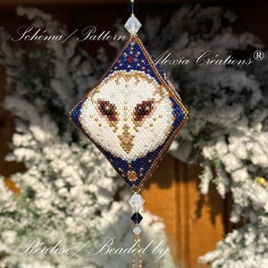 May include: A beaded owl ornament with a diamond shape, featuring a white face, brown eyes, and a navy blue border. The ornament is embellished with gold and clear beads, and has a beaded tassel. Text on the image reads "Schema / Pattern" and "Alexia Creations".