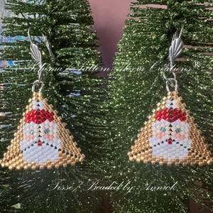 May include: A pair of triangular earrings featuring a beaded Santa Claus design. The Santa figure has a red hat, white beard, and peach-colored face, framed by metallic gold beads. Each earring hangs from a silver-toned metal ear wire hook.