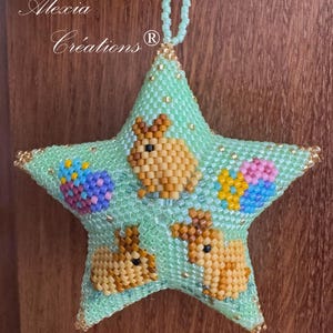 May include: A beaded star-shaped ornament in shades of green, gold, and brown, featuring Easter bunnies and eggs. The ornament has the text "Alexia Créations" and "Tissé / Beaded by Annick".