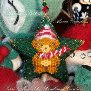 May include: A beaded Christmas ornament in the shape of a green star. The star features a teddy bear wearing a hat and scarf. The ornament is embellished with gold beads and a red and green crystal. The text "Alexia Créations" is visible.