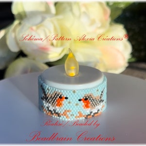 Puede incluir: Un portavelas de cuentas azul turquesa con dos petirrojos en la parte delantera. El portavelas está iluminado con una pequeña llama amarilla.