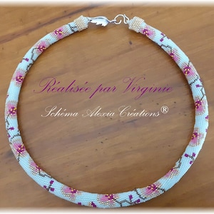 Peut inclure: Un collier de perles bleu clair avec un motif floral. Le collier est fait de petites perles et présente des accents roses et dorés. Le texte "Réalisée par Virginie" et "Schéma Alexia Creations®" est visible sur la surface en bois.