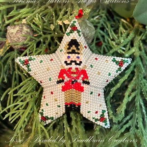 Puede incluir: Un adorno de estrella blanca con forma de estrella con un diseño de cascanueces rojo, dorado y negro. El adorno está adornado con cuentas blancas, rojas, verdes y doradas. El adorno está colgado de un árbol de Navidad verde.