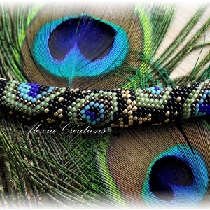 Peut inclure: Un bracelet en perles avec un motif géométrique noir, or, vert et bleu. Le bracelet est posé sur une plume de paon.