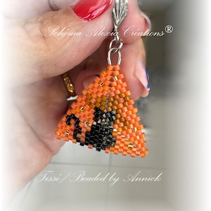 Könnte beinhalten: Orangefarbener und schwarzer dreieckiger Perlenohrring mit einem silbernen Haken. Der Ohrring ist mit einem schwarzen Katzenmotiv perlenbesetzt. Der Text "Schema Alexia Creations" ist auf dem Bild zu sehen.