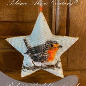 May include: A blue star-shaped beaded ornament featuring a robin perched on a branch. The ornament is decorated with gold beads and has a white background. The text "Schéma Alexia Creations®" is visible at the top of the ornament.