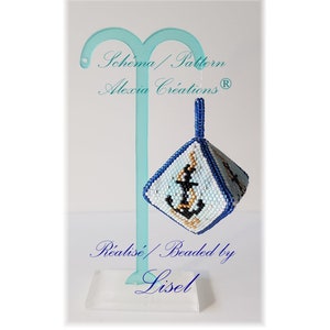 May include: A blue and white beaded ornament shaped like a cube with an anchor design. The ornament is hanging from a blue stand with the text "Schema/Pattern Alexia Creations (R)" and "Realise/Beaded by Lisel" on it.