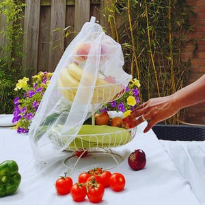 Könnte beinhalten: Ein weißer, dreistöckiger Obstkorb mit weißem Schleier, gefüllt mit Bananen, Äpfeln und anderem Obst. Der Korb steht auf einer weißen Tischdecke, umgeben von frischen Tomaten, Zwiebeln und einer grünen Paprika. Im Hintergrund Blumen und Grün.