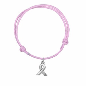 Puede incluir: Pulsera de cuerda rosa con un dije de cinta plateada. La pulsera está atada con un nudo y el dije cuelga del nudo.