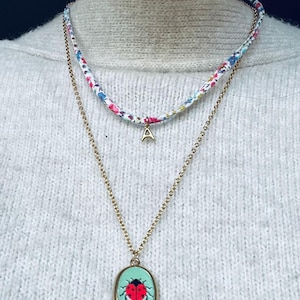 Peut inclure: Un collier superposé composé d'un ras du cou en tissu floral avec un pendentif initial doré et d'une chaîne dorée plus longue. Le pendentif de la chaîne plus longue est ovale avec un motif coccinelle sur fond turquoise.