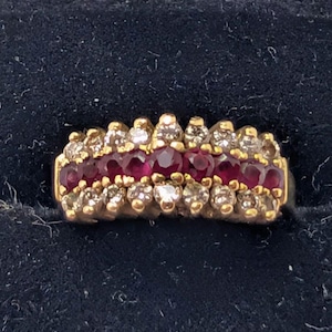 May include: A gold ring featuring a central horizontal row of seven round-cut deep red rubies flanked by two parallel rows of small, clear, round-cut diamonds. The band is constructed from yellow gold, showcasing a multi-stone cluster design.