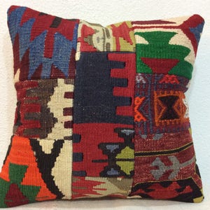 Puede incluir: Cojín cuadrado de patchwork con diversos patrones geométricos en tonos rojos, azules, verdes, naranjas y crema. El cojín tiene un aspecto texturizado y tejido, posiblemente hecho a mano o vintage.