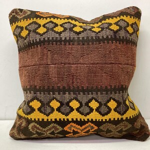May include: Square cushion with a woven design. The cushion features a central band of reddish-brown, bordered by bands of yellow, brown, and grey geometric patterns. The cushion's edges are a darker brown.