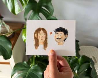 May include: A rectangular white card featuring a watercolor illustration of two people's heads facing each other, separated by a small orange heart. The person on the left has long brown hair, and the person on the right has short dark hair.