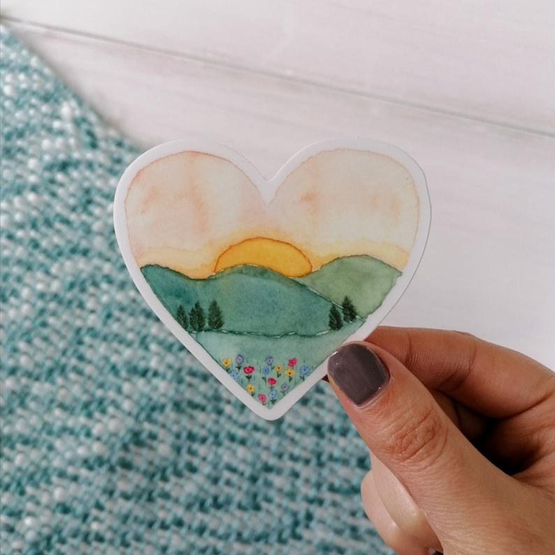 May include: A heart-shaped sticker with a watercolor painting of a sunset over rolling green hills with a field of colorful flowers in the foreground.