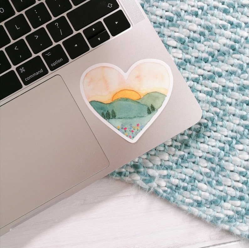 May include: A watercolor heart-shaped sticker with a sunset scene over a green mountain range. The sticker has a light orange, yellow, and green color scheme with a field of flowers at the bottom.