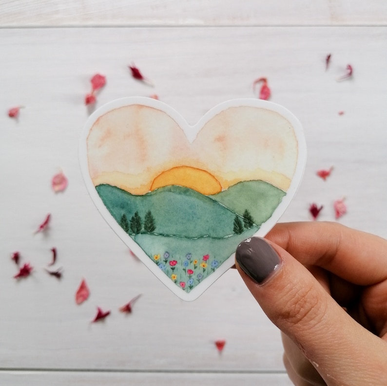 May include: A heart-shaped sticker with a watercolor painting of a sunset over rolling green hills and a field of wildflowers. The sun is a bright yellow and the sky is a soft orange and pink.