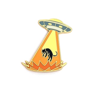 May include: A gold-colored enamel pin featuring a black cat being abducted by a UFO. The UFO is gray with a yellow light beam shining down on the cat. The cat is surrounded by orange flames.