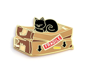 Puede incluir: Pin de esmalte dorado de un gato negro durmiendo encima de dos cajas de cartón con la etiqueta "FRAGILE".