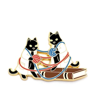 May include: Two black cats wearing white lab coats are playing with red and blue yarn on top of a brown book. The cats are facing each other and appear to be tangled in the yarn.