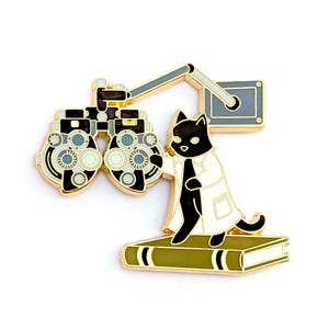 May include: Gold enamel pin featuring a black cat wearing a white lab coat and standing on a book. The cat is looking at an eye chart with a gold metal frame.