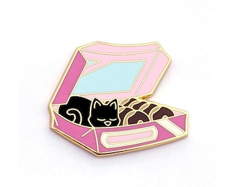 May include: A black cat sleeping in a pink and gold donut box with a blue and white lid. The cat is surrounded by donuts.