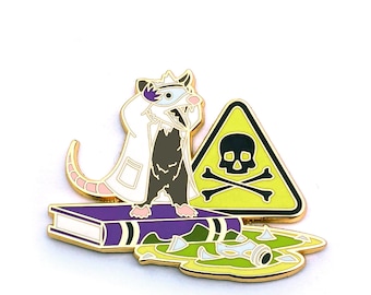 May include: A gold-colored enamel pin featuring a cartoon possum wearing a white lab coat and a purple eye patch, standing on a purple book with a yellow skull and crossbones warning sign behind it. The possum is standing on a puddle of green slime with shards of glass.