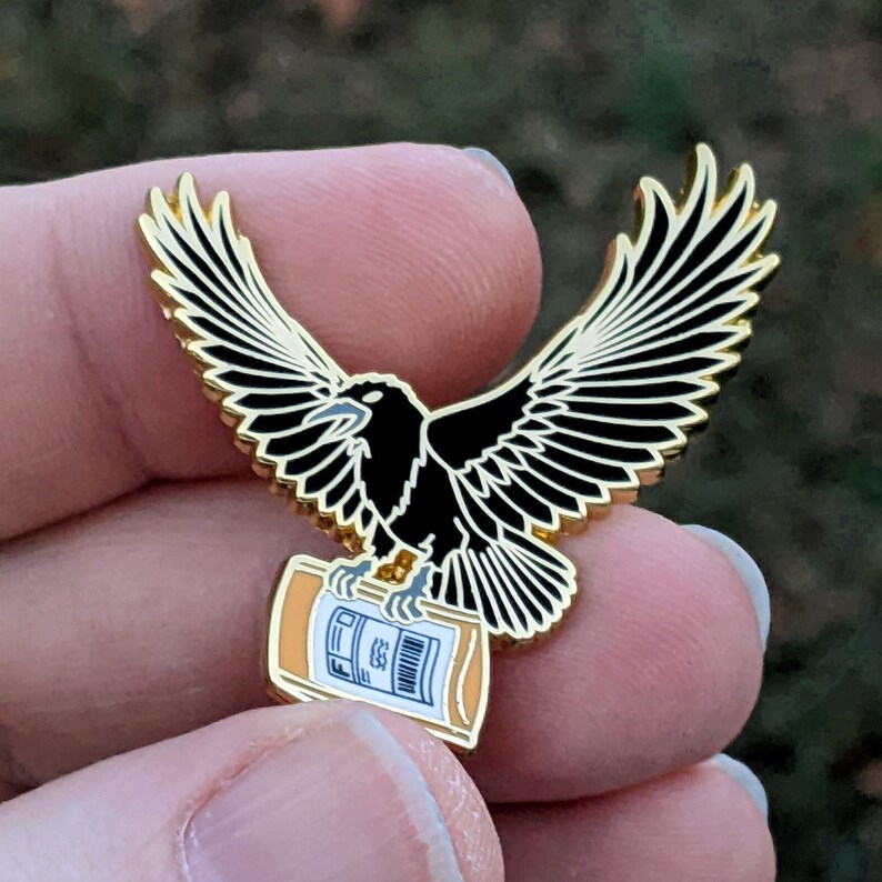 May include: Gold enamel pin featuring a black crow holding a small orange and white bottle with the text "FED" on it.