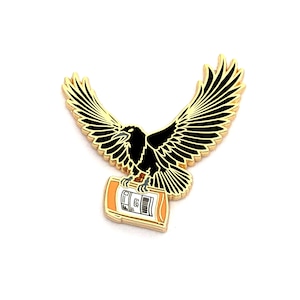 May include: A gold-colored pin featuring a black crow carrying a small orange bottle with a white label. The crow has its wings spread out.