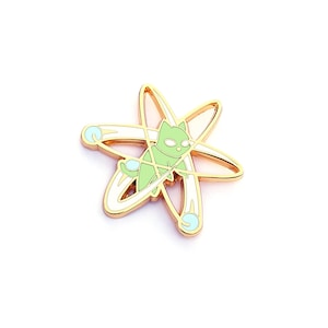 May include: A gold-colored metal pin featuring a light green cat inside a white and gold atom symbol. The cat has large eyes and a tail. The atom symbol has light blue accents.