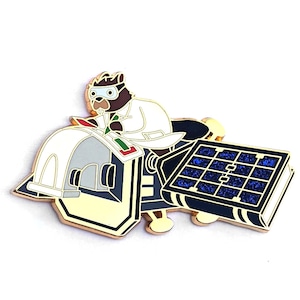 May include: A gold-colored enamel pin featuring a cartoon beaver wearing a lab coat and goggles, driving a small, white, and gold spacecraft with a blue solar panel.