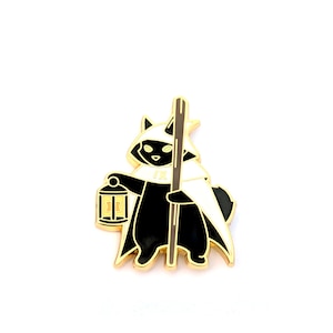 May include: A gold-colored enamel pin featuring a black cat dressed as a hooded figure, holding a lantern and a staff. The cat has a white cloak and a gold-colored trim. The pin is on a white background.