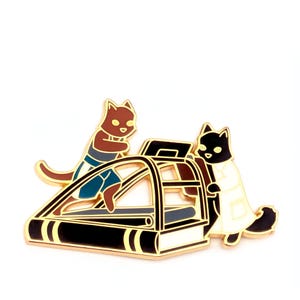 May include: A gold-toned metal enamel pin featuring two cats interacting with a treadmill. One brown cat wears blue shorts while running, and a black cat in a white lab coat stands beside the machine. The design has bold black outlines.