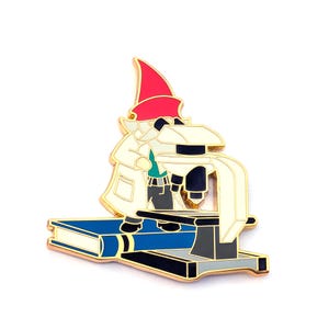 May include: An enamel pin depicting a gnome scientist in a white lab coat and red hat, using a microscope. The pin is set on a blue book with gold detailing, ideal for science lovers.