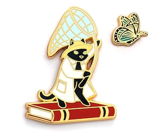 Pode incluir: Pin de esmalte dourado com um gato preto vestindo um jaleco branco de laboratório e segurando uma rede de borboleta. O gato está em pé em um livro vermelho com detalhes dourados. Uma borboleta verde e dourada está voando para longe do gato.
