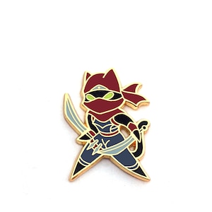 May include: A gold-colored enamel pin featuring a cartoon ninja cat character. The cat is wearing a maroon hood and mask, with green eyes. It holds two blue swords and is posed in a fighting stance. The pin has black, blue, and gold accents.