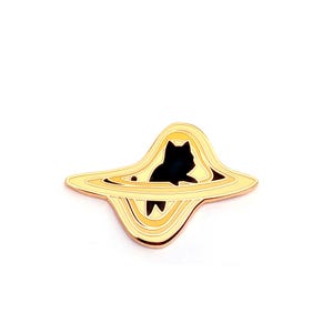 May include: A gold-toned metal enamel pin featuring a black silhouette of a cat positioned within a stylized, swirling yellow and gold ringed planet shape. The design includes concentric circular lines that create a sense of depth and movement.