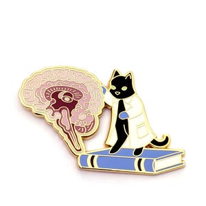 May include: A gold-toned enamel pin featuring a black cat wearing a white lab coat and blue gloves, standing on a blue book while touching a detailed anatomical cross-section of a human brain rendered in shades of pink, purple, and maroon.