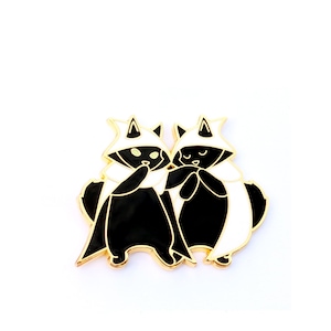 May include: A gold-toned enamel pin featuring two stylized black and white raccoon-like figures. Each figure wears a white cape and hat with a black triangle. The figures are close together, suggesting friendship or companionship.