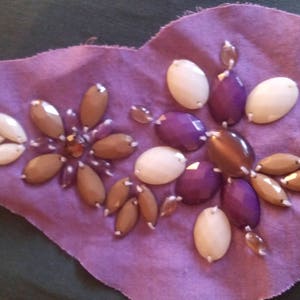 Peut inclure: Applique en tissu violet en forme de cœur, ornée de pierres précieuses. Les pierres sont blanches, marron et violettes, disposées en motif floral. L'applique est sur un fond vert foncé.