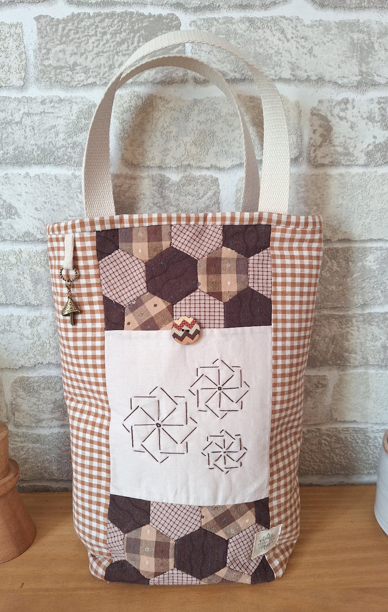 Peut inclure: Un sac cabas en vichy marron et blanc avec un motif patchwork et une poche blanche orn&eacute;e de motifs floraux brod&eacute;s. Le sac a un bouton marron et un petit charme.