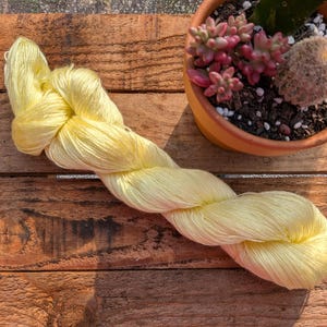 May include: A skein of pale yellow yarn is displayed on a weathered wooden surface. The yarn is twisted and coiled, showcasing its texture and color. A small terracotta pot with succulents and a cactus is in the background.