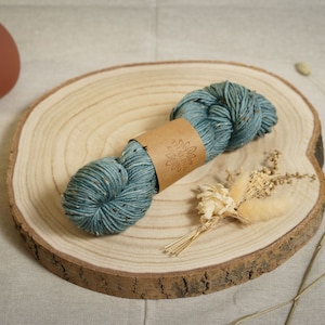 Può includere: Un gomitolo di filato blu turchese con macchie marroni e bianche, avvolto in carta marrone con un logo. Il filato è su una fetta di legno.