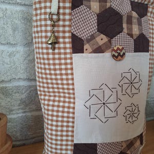 Peut inclure: Un sac cabas en vichy marron et blanc avec un motif patchwork. Le sac a une petite poche sur le devant avec une fermeture &agrave; bouton. Le sac a une bandouli&egrave;re en vichy marron et blanc avec un petit charme en forme de champignon.