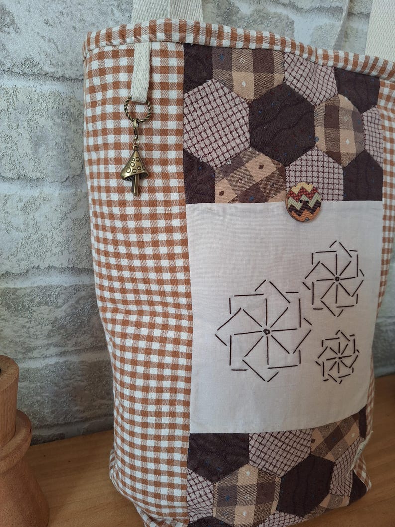 Peut inclure: Un sac cabas en vichy marron et blanc avec un motif patchwork. Le sac a une petite poche sur le devant avec une fermeture &agrave; bouton. Le sac a une bandouli&egrave;re en vichy marron et blanc avec un petit charme en forme de champignon.