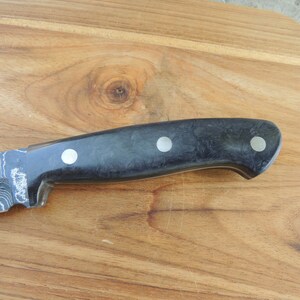 Puede incluir: Un cuchillo con patr&oacute;n negro y plateado con mango negro y tres detalles plateados. El cuchillo est&aacute; sobre una superficie de madera.