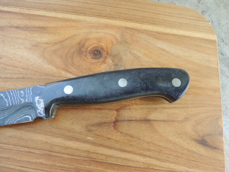 Puede incluir: Un cuchillo con patr&oacute;n negro y plateado con mango negro y tres detalles plateados. El cuchillo est&aacute; sobre una superficie de madera.