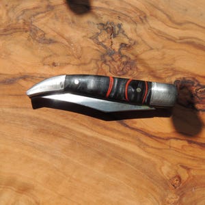 May include: A folding pocket knife with a curved silver metal blade and a black handle featuring two distinct red circular accents. The handle is secured with silver metal rivets at both ends and in the center, showcasing a sleek, compact design.