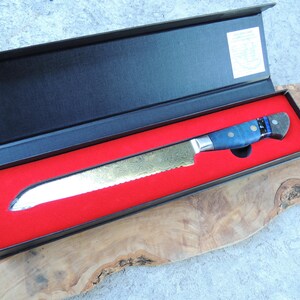 May include: A black box with a red interior, containing a silver bread knife with a blue and black handle. The knife has a serrated edge and a decorative pattern on the blade.