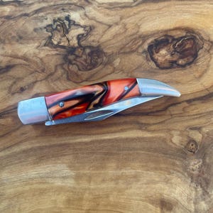 May include: A folding pocket knife with a curved, metallic blade and a handle featuring a marbled pattern of orange, black, and white swirls. The handle is secured with two silver-toned rivets and features a polished metal bolster at the end.